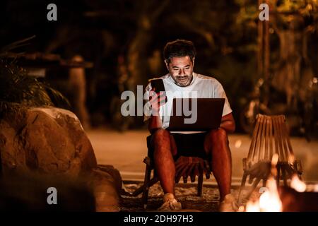 Smiling man with phone using laptop at night Stock Photo