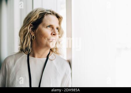 Mature entrepreneur looking away while leaning by window at workplace Stock Photo