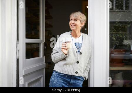 Smiling mature woman having coffee at entrance of art studio Stock Photo