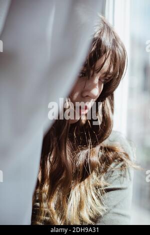 Close-up of young woman hiding behind window curtain Stock Photo