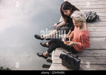 High angle view of happy businesswomen using smart phone while sitting on pier by lake Stock Photo
