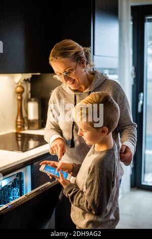 Mother guiding son while using mobile app over dishwasher in kitchen at home Stock Photo