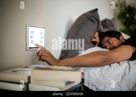 Man using digital tablet mounted on wall while lying on bed at smart home Stock Photo