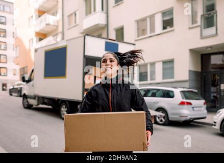 Confident young delivery woman carrying cardboard box on street in city Stock Photo