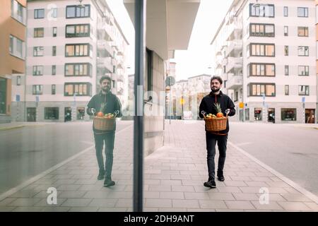 Full length of young delivery man carrying fruit basket on footpath in city Stock Photo