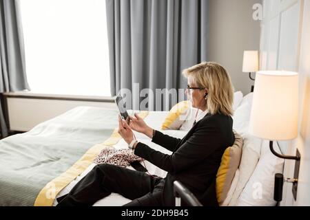 Side view of businesswoman using digital tablet sitting on bed at hotel room Stock Photo
