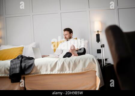 Businessman sitting on bed using laptop against wall in hotel room Stock Photo
