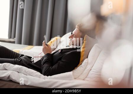 Side view of businessman listening music while lying on bed at hotel room Stock Photo