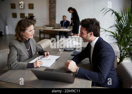 Confident senior female lawyer smiling while discussing over laptop with young businessman during meeting at office Stock Photo