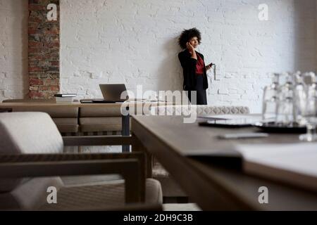 Confident young female lawyer talking on mobile phone while leaning on white wall at office Stock Photo