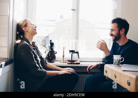 Happy couple sitting in kitchen against window at home Stock Photo