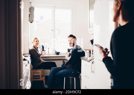 Happy couple sitting in kitchen while looking at female friend Stock Photo