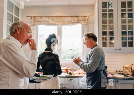 Man eating watermelon while friends talking in kitchen Stock Photo
