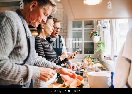 Senior man cutting watermelon by friends discussing over mobile phone in kitchen at home Stock Photo