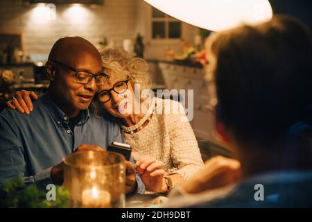 Bald senior sharing smart phone with woman while sitting at illuminated dining table Stock Photo
