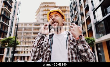 Angry builder, dissatisfied with deadline of work, swears at stress while talking to foreman on phone background of construction Stock Photo