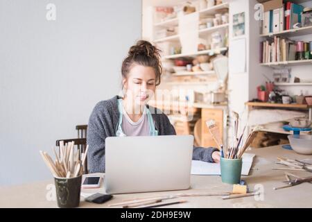 Young female potter using laptop while sitting at table in workshop Stock Photo