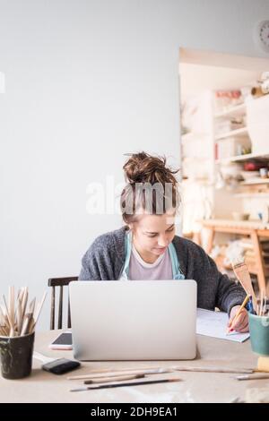 Young female potter writing on paper while sitting with laptop at table in workshop Stock Photo