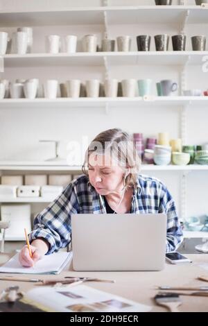 Mature female potter writing on paper while sitting with laptop at table in workshop Stock Photo