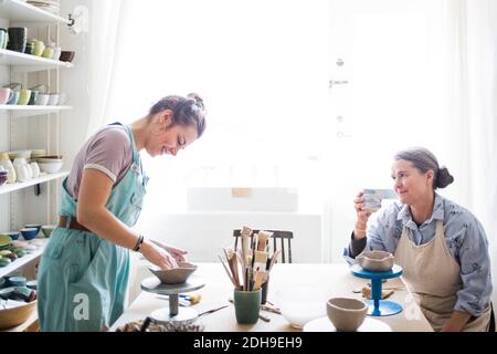 Senior female potter looking at young ceramic artist molding clay against window in workshop Stock Photo