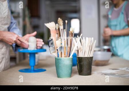 Hand tools and paintbrushes in containers on workbench with female potters working in background Stock Photo