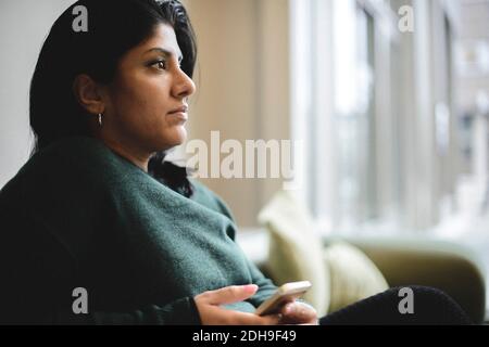 Thoughtful businesswoman holding smart phone while sitting at office lobby Stock Photo