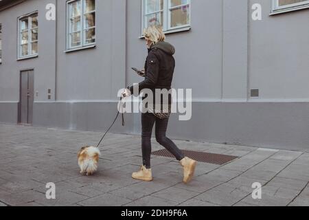 Full length of blond woman walking with Pomeranian on footpath by building in city Stock Photo