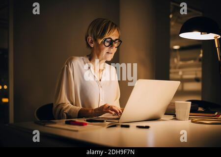 Determined businesswoman using laptop while working late in illuminated creative workplace Stock Photo