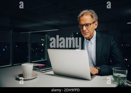 Confident senior businessman using laptop while sitting at illuminated desk in creative office Stock Photo