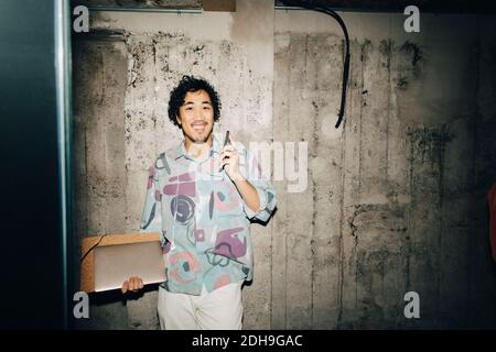 Portrait of smiling businessman with laptop standing against wall in creative office Stock Photo