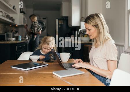 Woman using laptop while daughter having breakfast with family in background at home Stock Photo