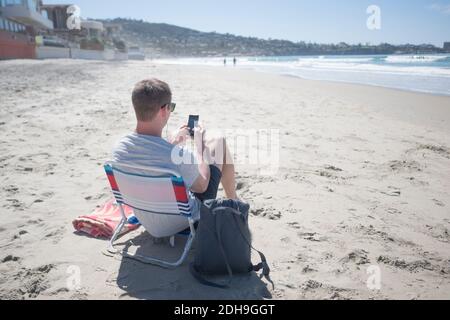 Rear view of man using smart phone while sitting on chair at beach Stock Photo