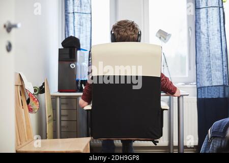 Rear view of teenage boy wearing headphones while sitting on chair at home Stock Photo