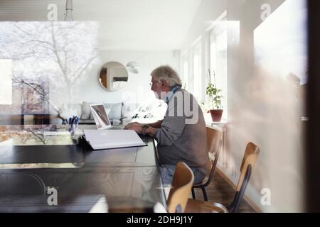 Senior man using laptop while sitting at table seen through window Stock Photo