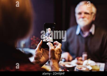 Senior woman photographing man through smart phone while having breakfast at table in hotel Stock Photo