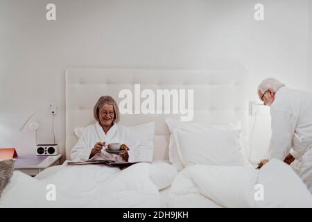 Happy senior woman having coffee while man standing by bed in hotel room Stock Photo