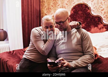 Gay couple looking at mobile phone while sitting on bed in hotel room Stock Photo