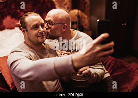 Man kissing boyfriend taking selfie while enjoying wine in hotel room Stock Photo