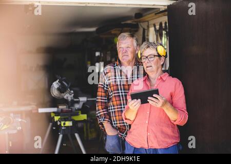 Thoughtful senior couple with digital tablet looking away while standing at workshop doorway Stock Photo