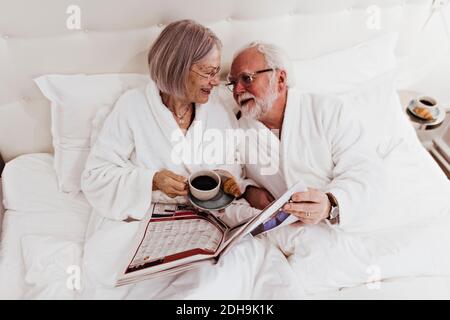 Senior woman looking at man holding newspaper while having coffee on bed in hotel Stock Photo