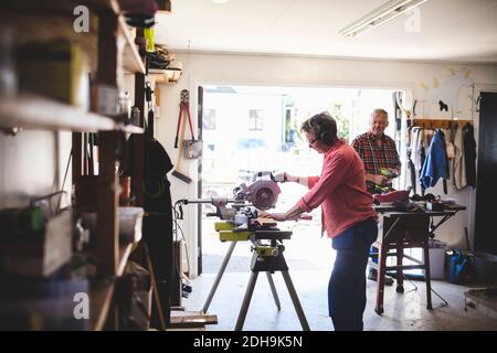 Side view of senior woman using circular saw while man holding drill in background Stock Photo