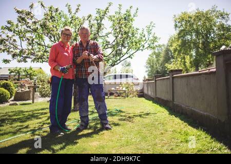 Full length portrait of senior couple holding hammer and garden hose while standing at yard Stock Photo