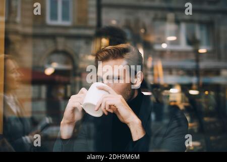 Businessman drinking coffee while looking away in cafe seen through glass window Stock Photo