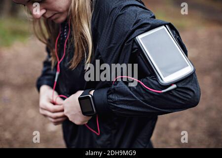 Midsection of female athlete with smart phone in arm band fixing headphones to jacket in forest Stock Photo