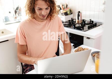 Caucasian handsome pleased guy using laptop while having breakfast at kitchen Stock Photo