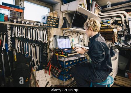 Side view of female mechanic holding disposable cup while using laptop at auto repair shop Stock Photo