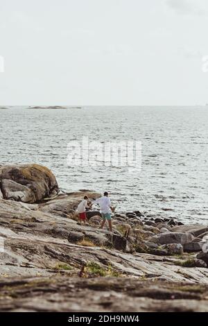 Rear view of father with son walking towards sea on rocky land against sky Stock Photo