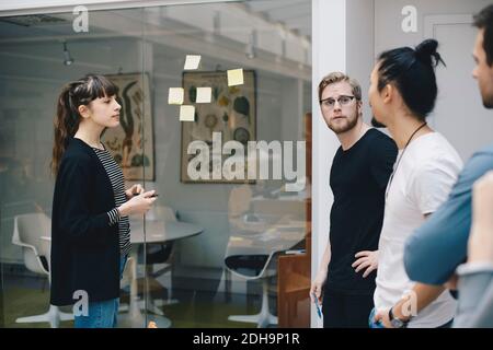 Male and female programmers discussing over adhesive notes stuck on glass in office Stock Photo