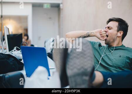 Tired computer programmer yawning while sitting with feet up at desk in office Stock Photo