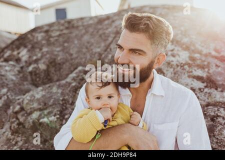 Portrait of baby girl with father looking away while sitting on rock formation Stock Photo